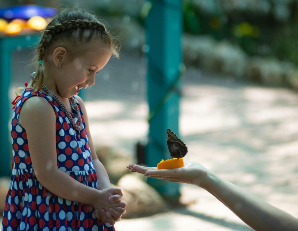 A young girl joyfully watches a butterfly perched on a hand outdoors.