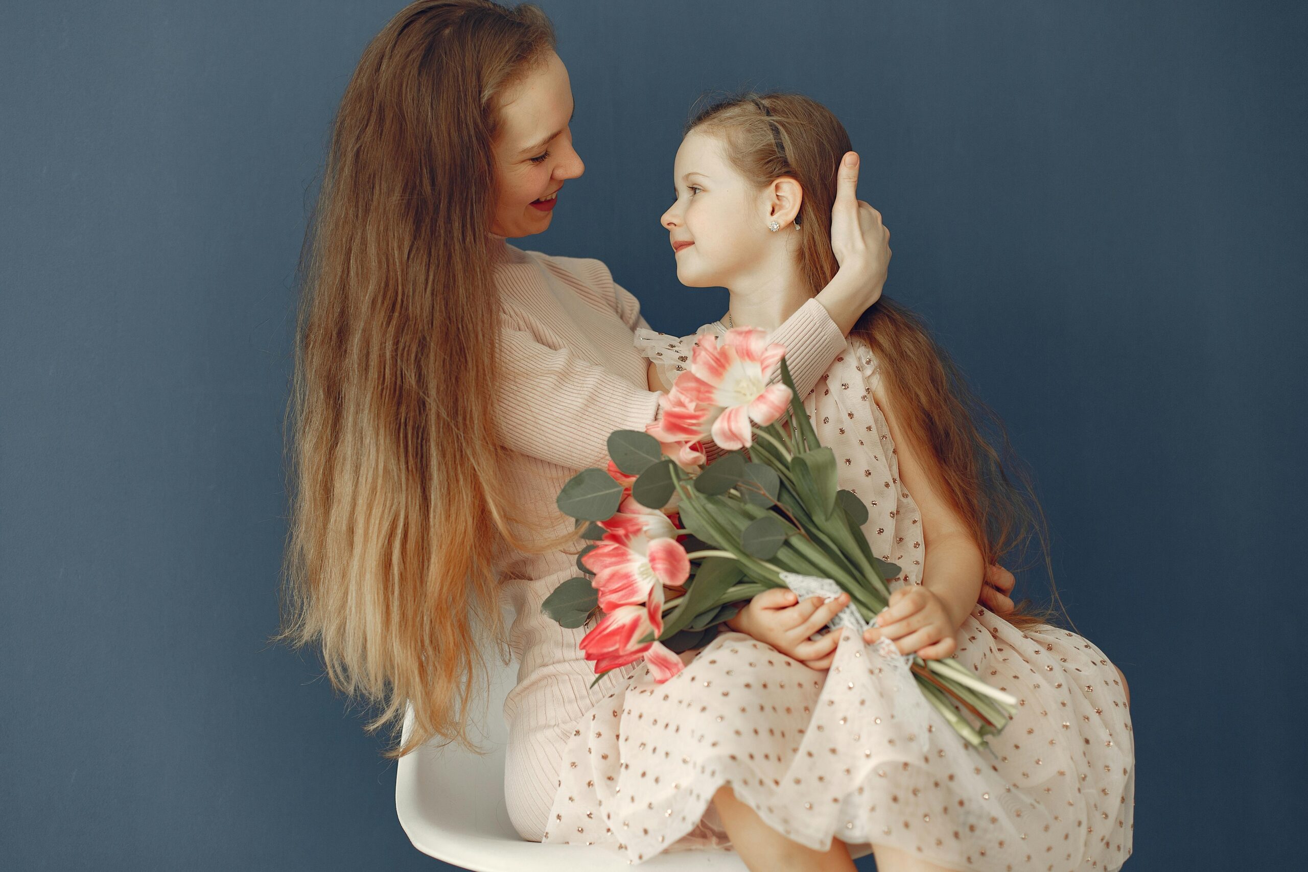 A touching moment of a mother embracing her daughter with a bouquet of pink flowers against a blue background.