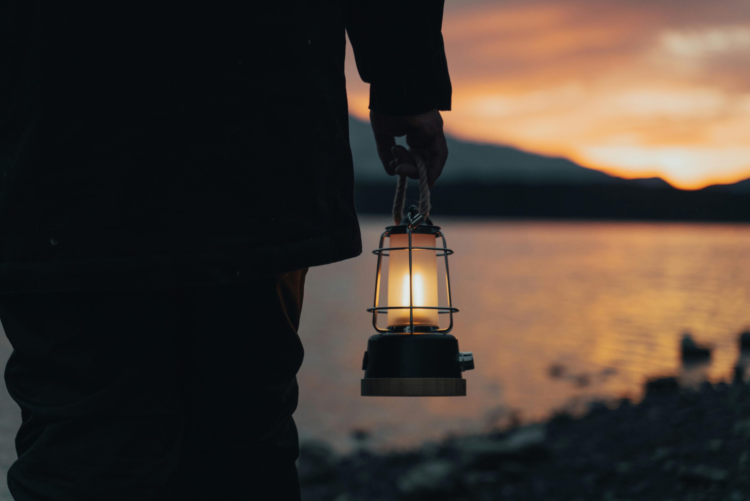 A person holding a glowing lantern beside a tranquil lake during sunset, creating a moody and calming scene.